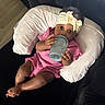 baby, infant, bottle, headband, pink_clothing, cushion, sofa, indoor, child, cute, relaxing, looking_at_camera, comfortable, small_hands, foot, dark_furniture, portrait, human, toddler, feeding