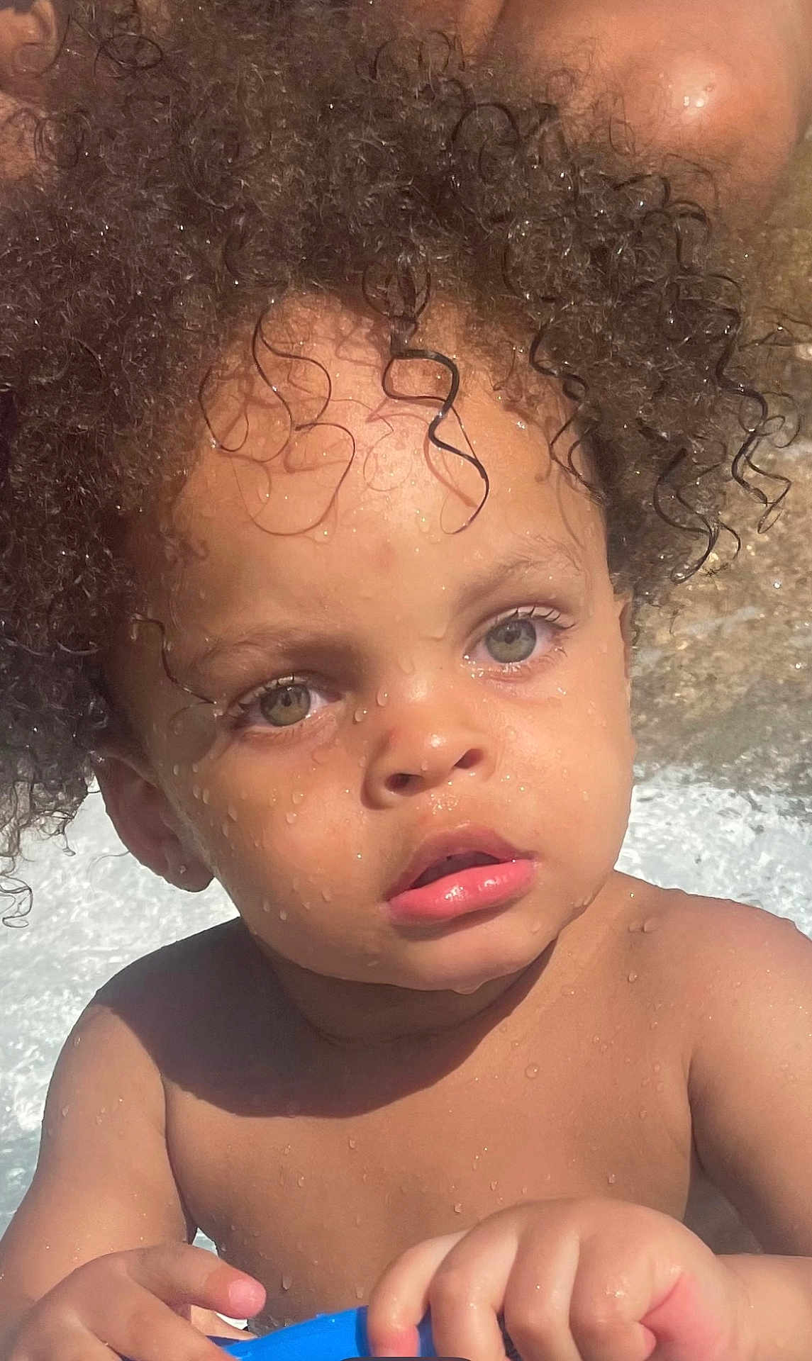 Sulieman joined the competition — help win amazing prizes! child, toddler, curly_hair, wet_skin, water_droplets, blue_object, outdoor, sunlight, face, eyes, hands, playing, summer, skin, portrait, expression, closeup, baby, person, nature