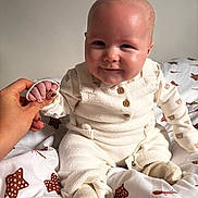 Vinnie is registered to the contest to win money with this photo: baby, bed, blanket, child, cozy, cute, hand_holding, happy, indoor, infant, knitwear, pattern, person, portrait, sitting, smile, soft_light, star_pattern, warm, white_clothing