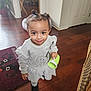 toddler, child, smiling, indoors, floor, wooden_floor, sippy_cup, hair_buns, white_dress, black_boots, rug, door, plant, household, person, young_child, cute, happy, standing, girl