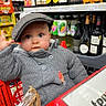 baby, child, shopping_cart, hat, grey_sweater, store, grocery_store, bottles, beverages, shelves, curious, face, person, indoor, retail, shopping, cute, infant, cap, hand_raised