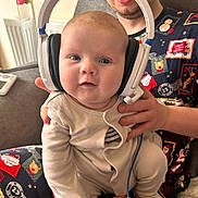Teagan is registered to the contest to win money with this photo: baby, headphones, person, face, glasses, pajamas, christmas_theme, couch, hand, infant, indoor, cute, child, smile, seated, clothing, cozy, holiday, adult, parent