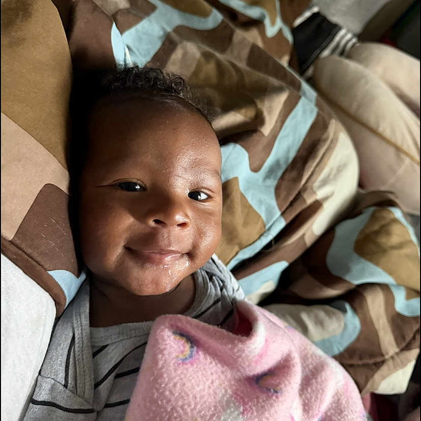 Noah is registered to the contest to win money with this photo: baby, blanket, bedspread, smile, infant, striped_clothing, face, indoors, child, comfort, patterned, fabric, person, cute, lying_down, soft_light, happy, young_child, cozy, portrait