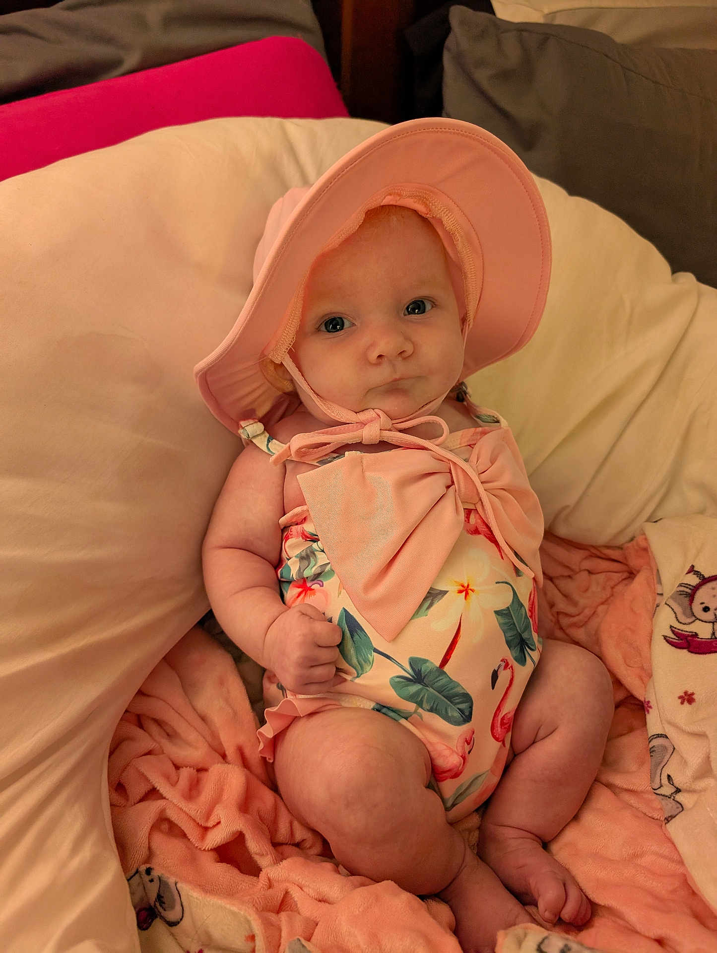 Adeline joined the competition — help win amazing prizes! baby, infant, pink_hat, floral_swimsuit, bow, blanket, soft_fabric, couch, indoors, cute, chubby_arms, chubby_legs, sitting, face, headwear, summer_clothing, child, person, portrait, skin