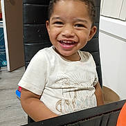 Elijah is registered to the contest to win money with this photo: child, toddler, smiling, curly_hair, white_tshirt, black_chair, indoor, door, table, happy, person, face, young, cute, seated, wood_floor, background, light, home, casual