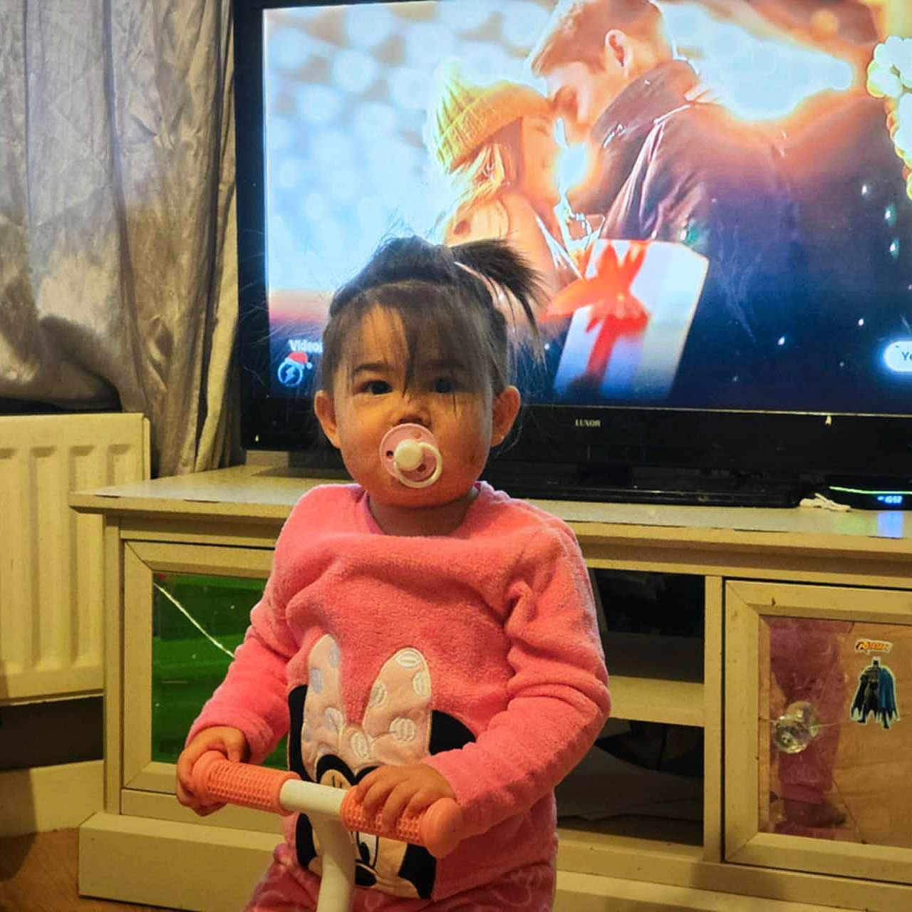 Harley-Mae is registered to the contest to win money with this photo: child, cozy, curtains, cute, floor, furniture, home, indoor, living_room, minnie_mouse, pacifier, person, pink_sweater, radiator, reflection, sticker, television, toddler, toy_scooter, young_child