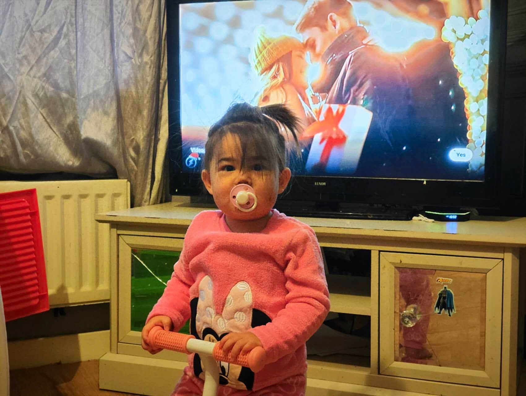 Harley-Mae is registered to the contest to win money with this photo: child, toddler, pacifier, pink_sweater, minnie_mouse, toy_scooter, indoor, living_room, television, furniture, curtains, radiator, floor, reflection, sticker, person, young_child, cute, home, cozy