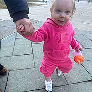 Layla is registered to the contest to win money with this photo: adult_hand, bow, building, candid, child, city, hair, hand_holding, outdoor, pavement, pink_hoodie, playful, portrait, sidewalk, sippy_cup, smile, sneakers, toddler, urban, walking