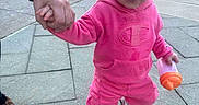Layla is registered to the contest to win money with this photo: adult_hand, bow, building, candid, child, city, hair, hand_holding, outdoor, pavement, pink_hoodie, playful, portrait, sidewalk, sippy_cup, smile, sneakers, toddler, urban, walking