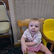 Layla is registered to the contest to win money with this photo: toddler, child, wooden_chair, yellow_chair, striped_shirt, pink_shorts, carpet, patterned_wall, indoor, furniture, person, sitting, barefoot, curious, baby, floor, metal_chair, hand, face, blue_eyes