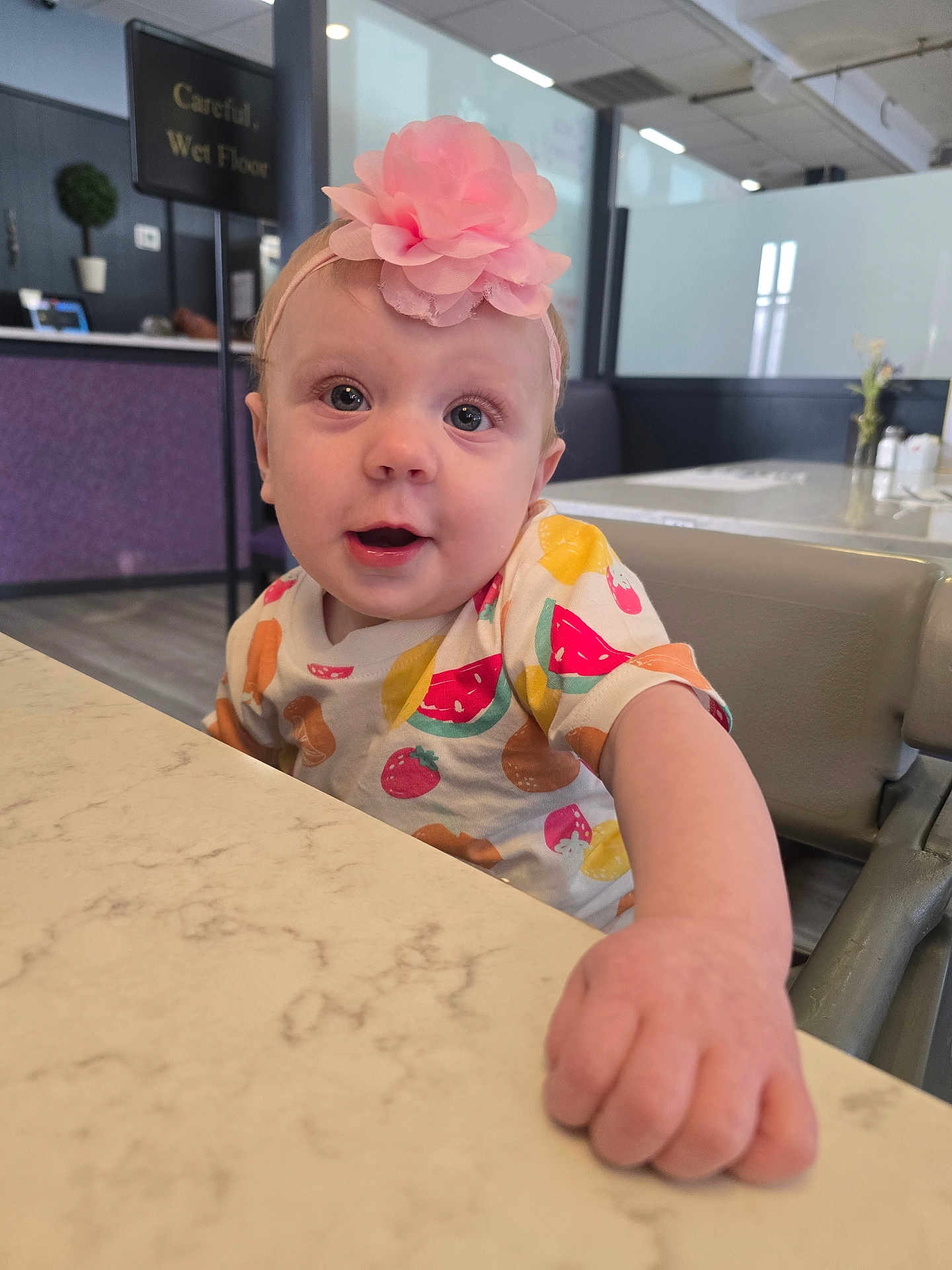 Layla joined the competition — help win amazing prizes! baby, child, infant, headband, flower, high_chair, table, fruit_pattern, shirt, indoor, cafe, face, hand, curious, expression, furniture, background, sign, marble_table, person
