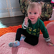 Layla is registered to the contest to win money with this photo: child, toddler, girl, pigtails, green_shirt, socks, sitting, pink_mat, orange_rug, pacifier_ring, toy, indoor, living_room, sofa, blanket, baby_gate, hair_tie, smile, portrait, floor