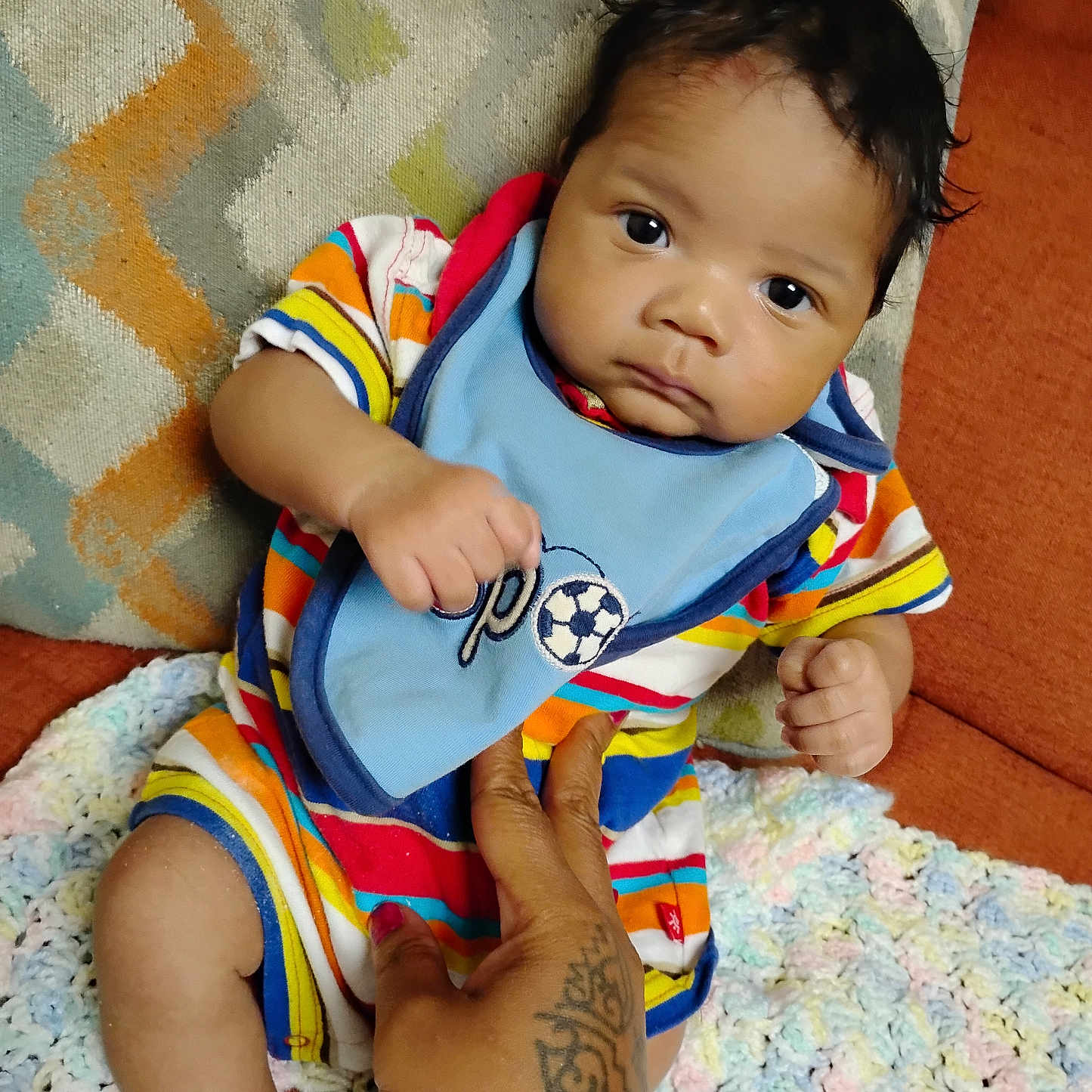 Josiah joined the competition — help win amazing prizes! baby, bib, child, crocheted_blanket, cushion, cute, expression, hand, indoor, infant, looking_at_camera, patterned_fabric, person, portrait, seated, skin, sofa, striped_clothing, tattoo, young_child