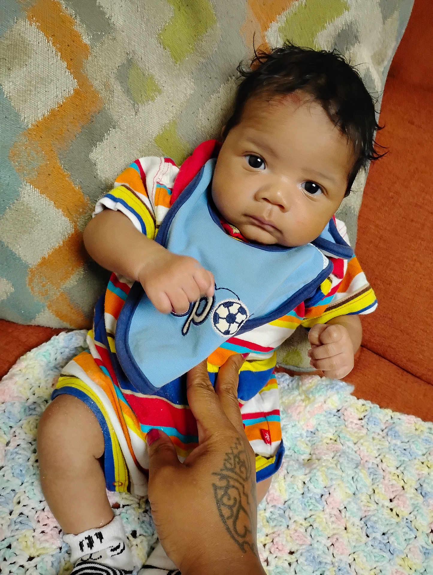 Josiah joined the competition — help win amazing prizes! baby, infant, child, hand, tattoo, bib, striped_clothing, crocheted_blanket, cushion, sofa, patterned_fabric, indoor, person, cute, portrait, seated, young_child, looking_at_camera, skin, expression