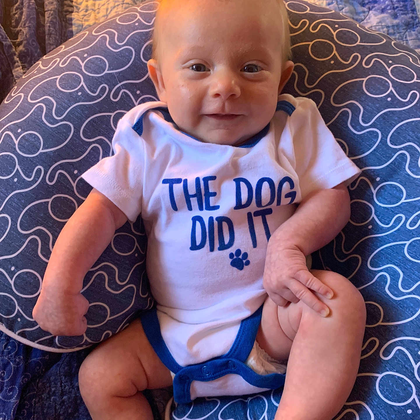 Dayton is registered to the contest to win money with this photo: baby, blue, child, cushion, foot, hand, happy, indoor, infant, leg, mohawk_hairstyle, onesie, patterned_textile, person, playful, quilt, skin, smiling, text_on_clothing, white