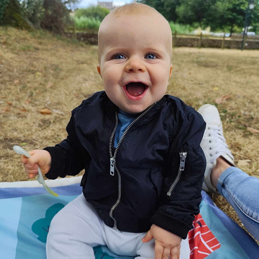 Liam participe au concours pour gagner de l'argent avec cette photo : baby, blanket, blue_eyes, casual, child, daylight, footwear, fork, grass, happy, jacket, joy, nature, outdoor, pants, park, person, sitting, smiling, trees