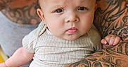 Enoch is registered to the contest to win money with this photo: baby, child, infant, portrait, face, eyes, looking_at_camera, sitting, onesie, clothing, tattoos, adult, arms, holder, indoor, close_up, photography, head, skin, family