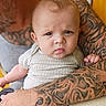 baby, child, infant, portrait, face, eyes, looking_at_camera, sitting, onesie, clothing, tattoos, adult, arms, holder, indoor, close_up, photography, head, skin, family
