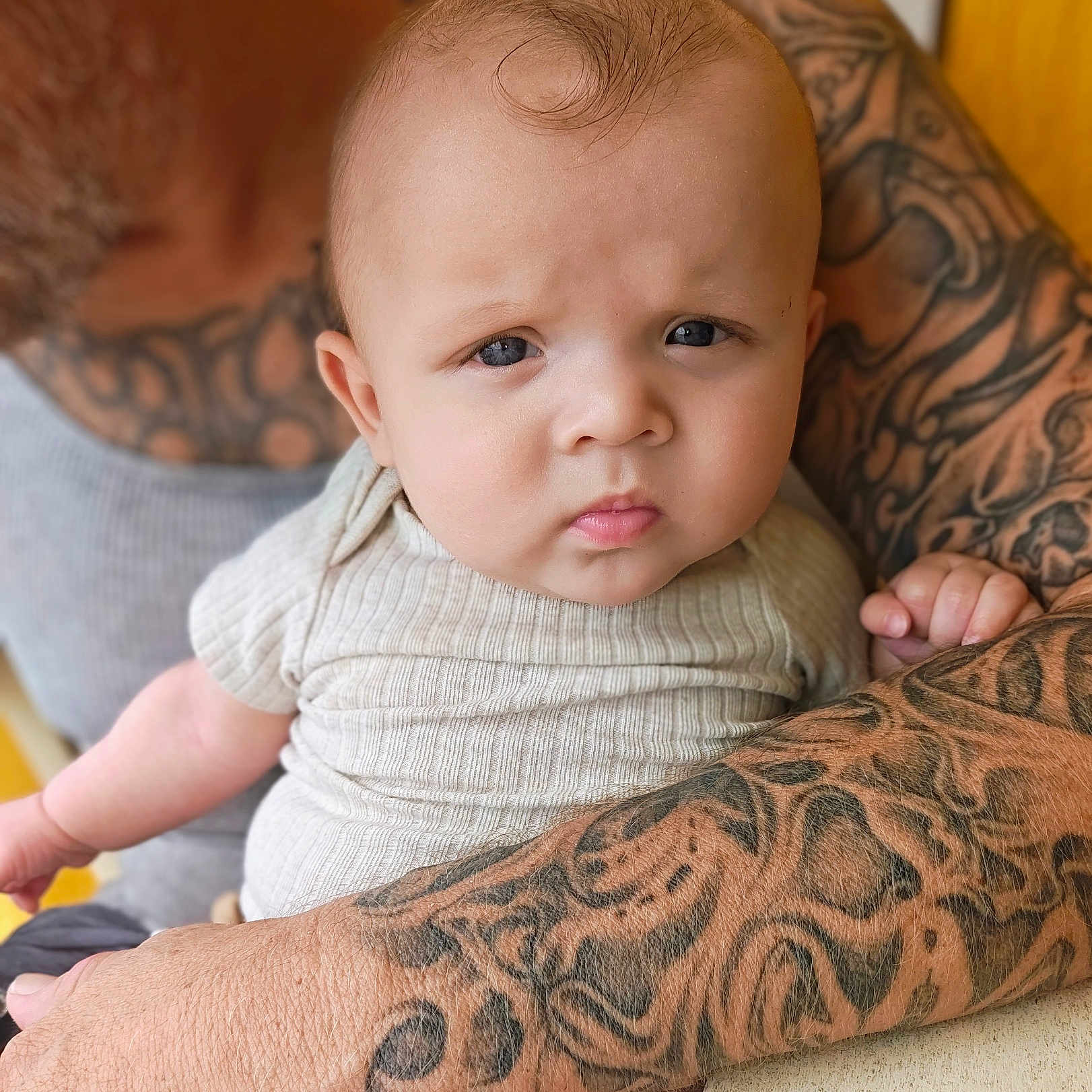 Enoch is registered to the contest to win money with this photo: baby, child, infant, portrait, face, eyes, looking_at_camera, sitting, onesie, clothing, tattoos, adult, arms, holder, indoor, close_up, photography, head, skin, family