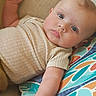 baby, infant, child, portrait, lying_down, wide_eyed, blue_eyes, raised_hand, smiling, face, head, onesie, clothing, cushion, patterned_fabric, indoor, close_up, relaxed, photography, colorful_background