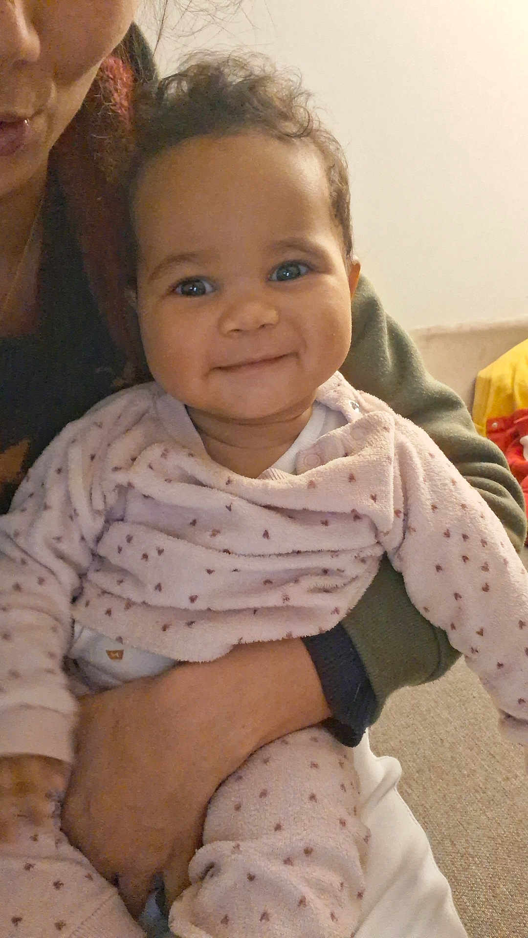 Aliyah a rejoint le concours — aidez-le/la à gagner de superbes lots ! baby, child, smiling, curly_hair, pink_outfit, heart_pattern, adult_hand, indoor, cozy, cute, portrait, happy, person, holding, closeup, infant, warm_lighting, family, love, casual