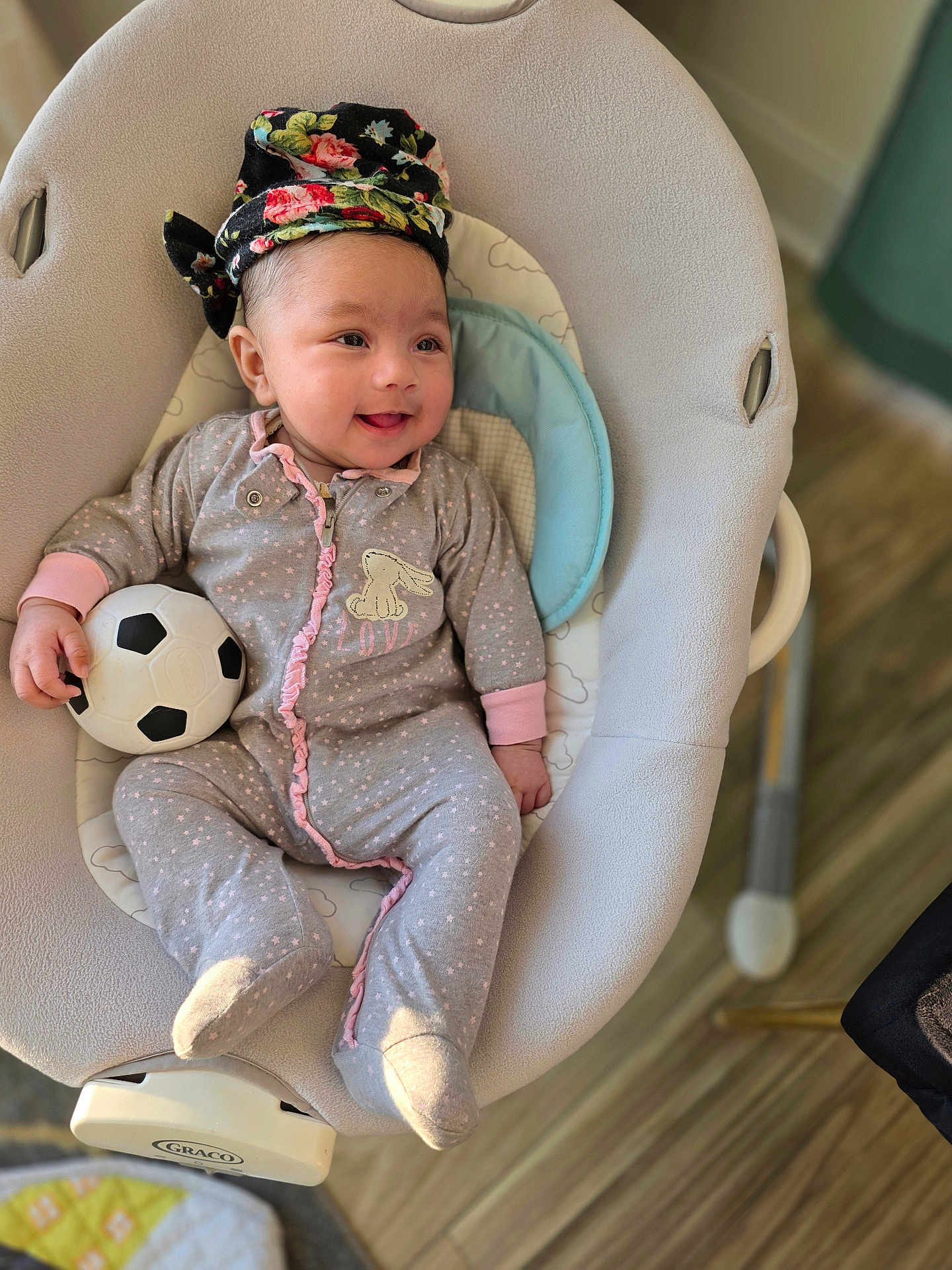 Vedanya Aruna Jain is registered to the contest to win money with this photo: baby, infant, smiling, onesie, headwrap, soccer_ball, baby_rocker, indoor, flooring, wood_floor, cozy, child, cute, pink, grey_clothing, toy, happy, playful, face, person