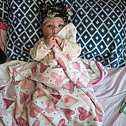 Vedanya Aruna Jain is registered to the contest to win money with this photo: baby, infant, blanket, heart_pattern, bed, pillow, blue_pillow, diamond_pattern, headwrap, floral_headwrap, hand, fingers, clothing, cute, lying_down, indoors, person, face, wide_eyes, curious