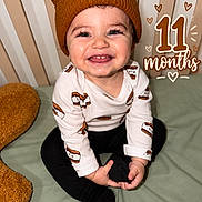 Hunter joined the competition — help win amazing prizes! baby, child, infant, smiling, happy, portrait, sitting, bed, bedroom, knit_hat, bear_hat, long_sleeves, black_pants, clothing, hands, face, teeth, striped_background, milestone, text_overlay