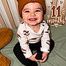 baby, child, infant, smiling, happy, portrait, sitting, bed, bedroom, knit_hat, bear_hat, long_sleeves, black_pants, clothing, hands, face, teeth, striped_background, milestone, text_overlay