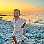 child, beach, sand, sunset, sea, water, sky, barefoot, smiling, playful, running, light_clothing, outdoor, nature, vacation, sunlight, horizon, shore, fun, happy