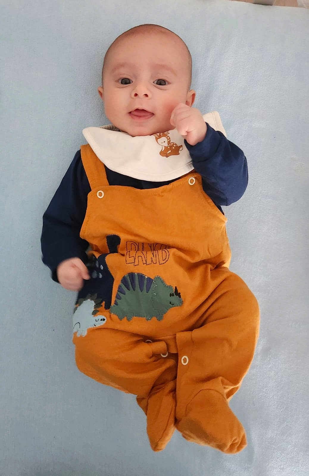 Arthur a rejoint le concours — aidez-le/la à gagner de superbes lots ! baby, infant, dinosaur_outfit, orange_clothing, blue_blanket, cute, smiling, lying_down, young_child, playful, face, person, portrait, indoors, cozy, hand_up, expression, soft_surface, child, adorable