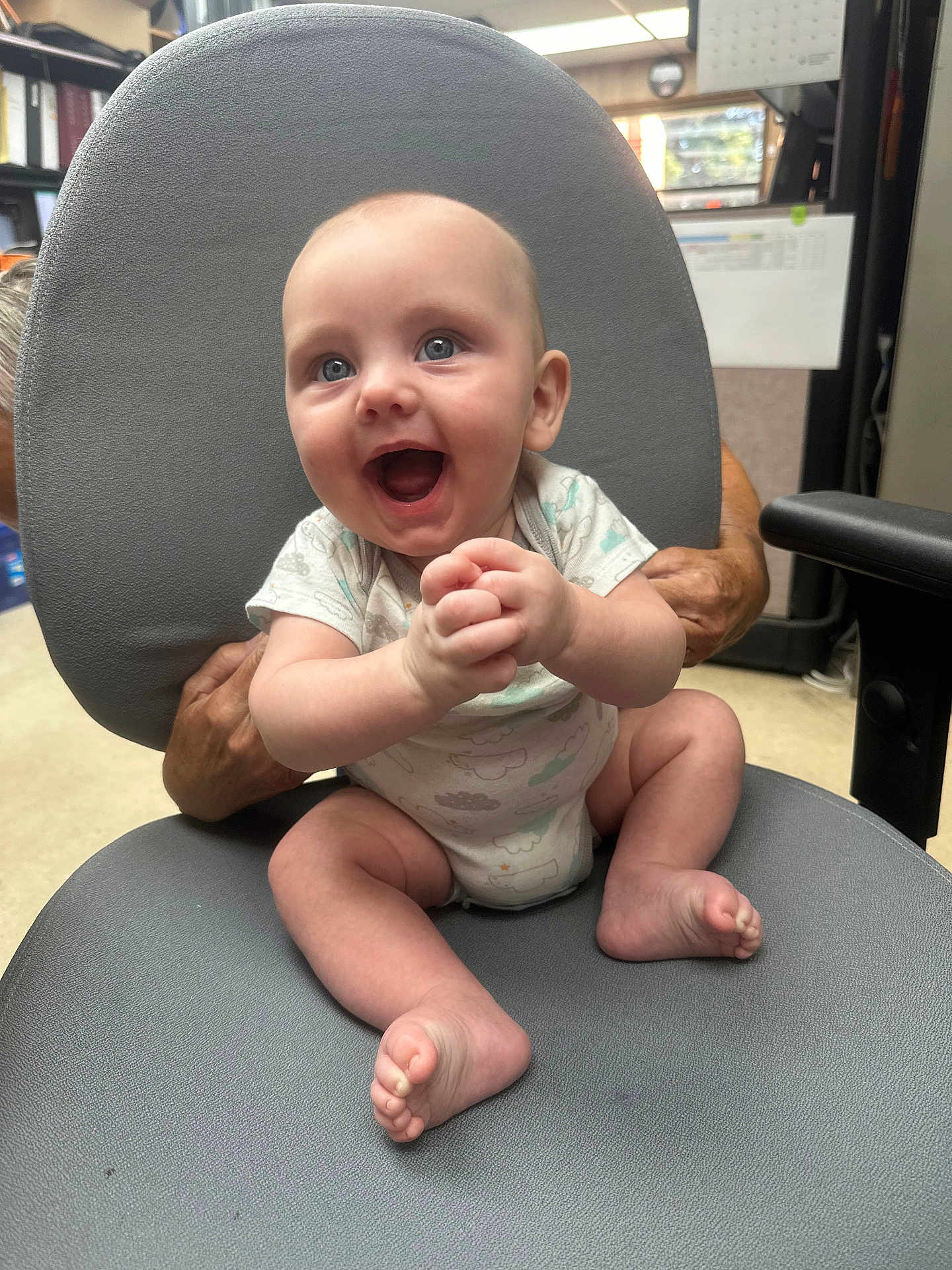 Jaxon is registered to the contest to win money with this photo: baby, infant, smiling, happy, hands, feet, clothing, onesie, chair, office, adult_hands, supporting, indoor, person, face, blue_eyes, skin, seated, expression, joyful