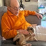 Bluey Wilson is registered to the contest to win money with this photo: casual_clothing, couch, cozy, elderly_man, framed_picture, gentle, glasses, gray_furniture, hand, home, indoor, lamp, orange_jacket, pet, pet_cage, puppy, relaxing, seated, watch, white_pants
