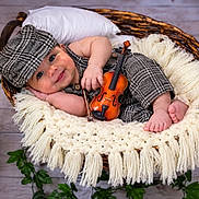 Taim is registered to the contest to win money with this photo: baby, infant, child, portrait, smiling, violin, musical_instrument, basket, blanket, knitwear, hat, cap, sitting, lying_down, indoor, studio, photography, greenery, vintage_clock, cute