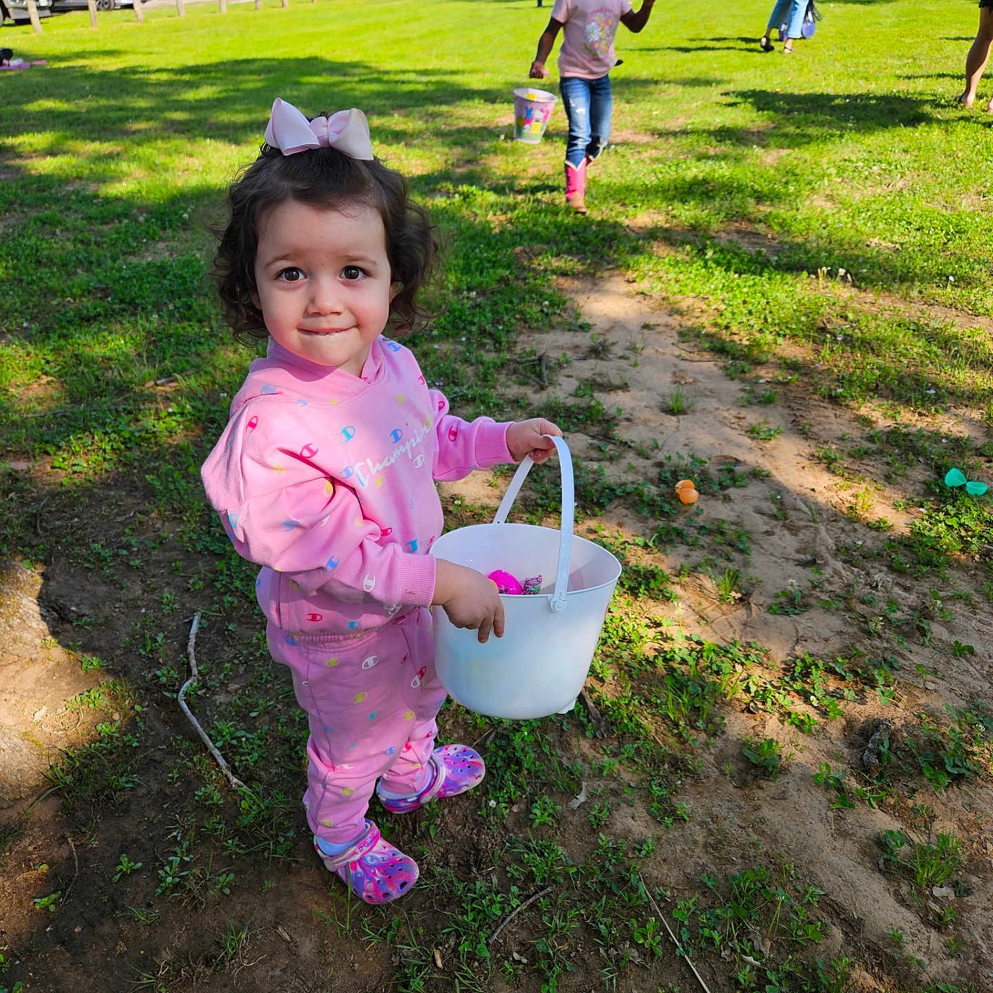 Estela is registered to the contest to win money with this photo: activity, boots, bow, bucket, child, daytime, easter_eggs, fun, grass, jeans, nature, outdoor, park, people, pink_clothing, play, smile, sunlight, toddler, vehicles