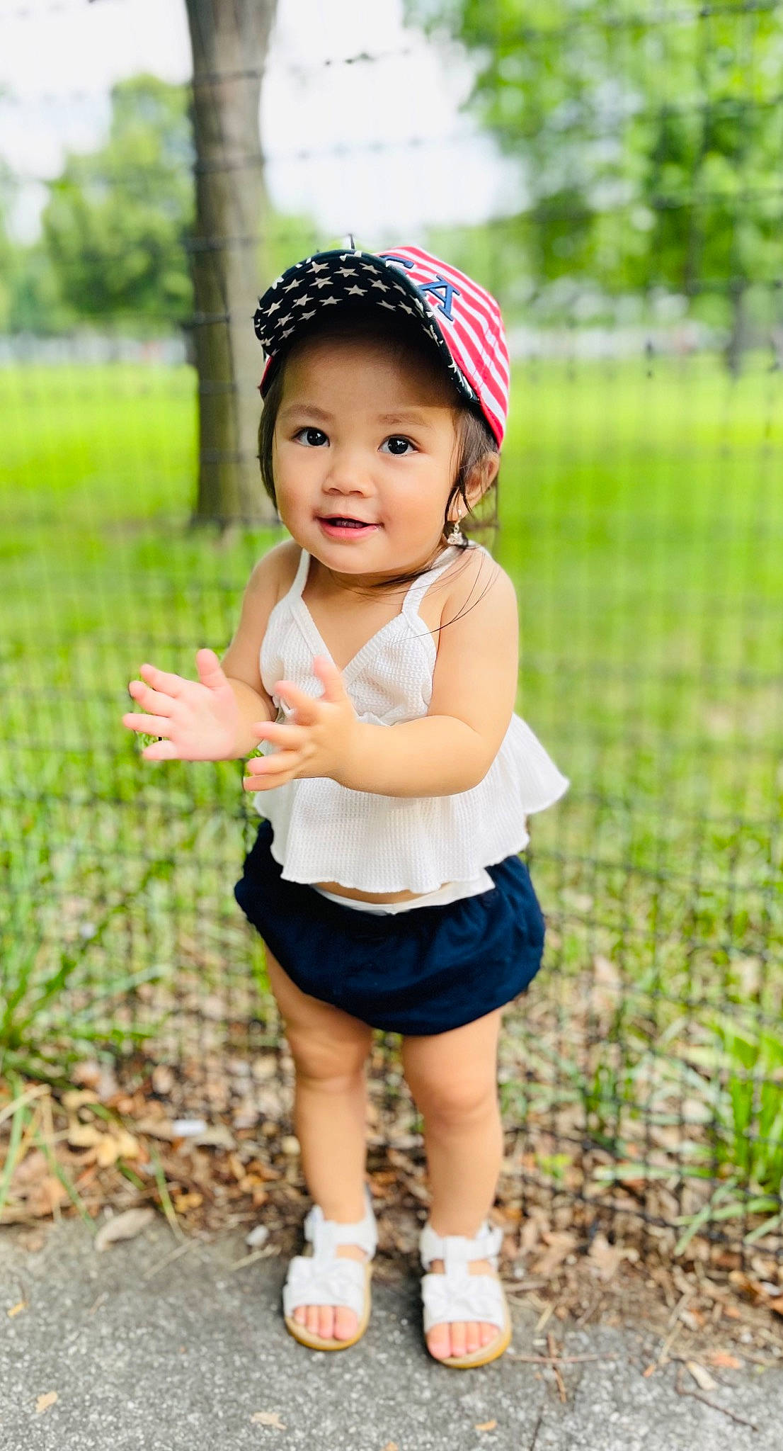 Amelia is registered to the contest to win money with this photo: baby, baby_toddler_clothing, cap, child, eye, face, fence, flash_photography, grass, happy, head, headband, headgear, headwear, joy, leisure, pattern, people_in_nature, person, plant