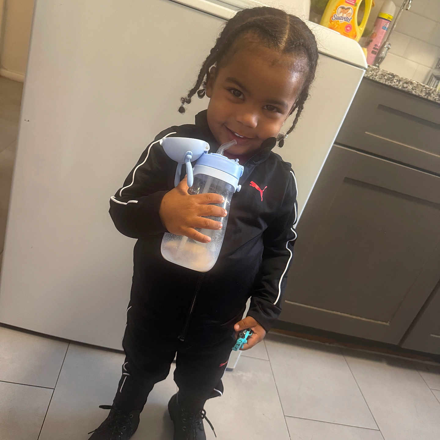 Jordan is registered to the contest to win money with this photo: child, smiling, braided_hair, black_tracksuit, puma_logo, water_bottle, toy_car, kitchen, washing_machine, laundry_detergent, floor_tiles, cabinet, countertop, young_child, indoors, happy, standing, black_boots, playful, portrait