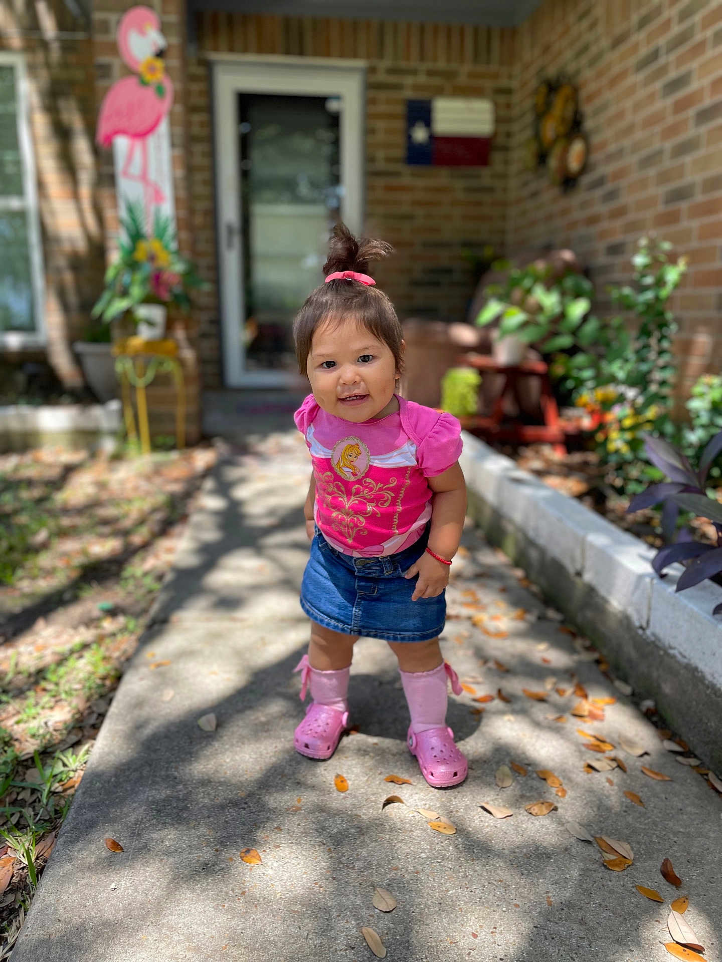 Marina Padilla is registered to the contest to win money with this photo: toddler, child, smiling, outdoor, sidewalk, pink_clothing, denim_skirt, pink_crocs, sunlight, brick_wall, plants, flower_pot, door, flamingo_decor, greenery, shadow, person, cute, happy, portrait
