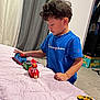 Ignacio joined the competition — help win amazing prizes! child, toy_train, blue_shirt, bedspread, indoor, carpet, curtains, toy_car, playing, young_child, person, furniture, room, clothing, hair, face, hand, floor, leisure, concentration