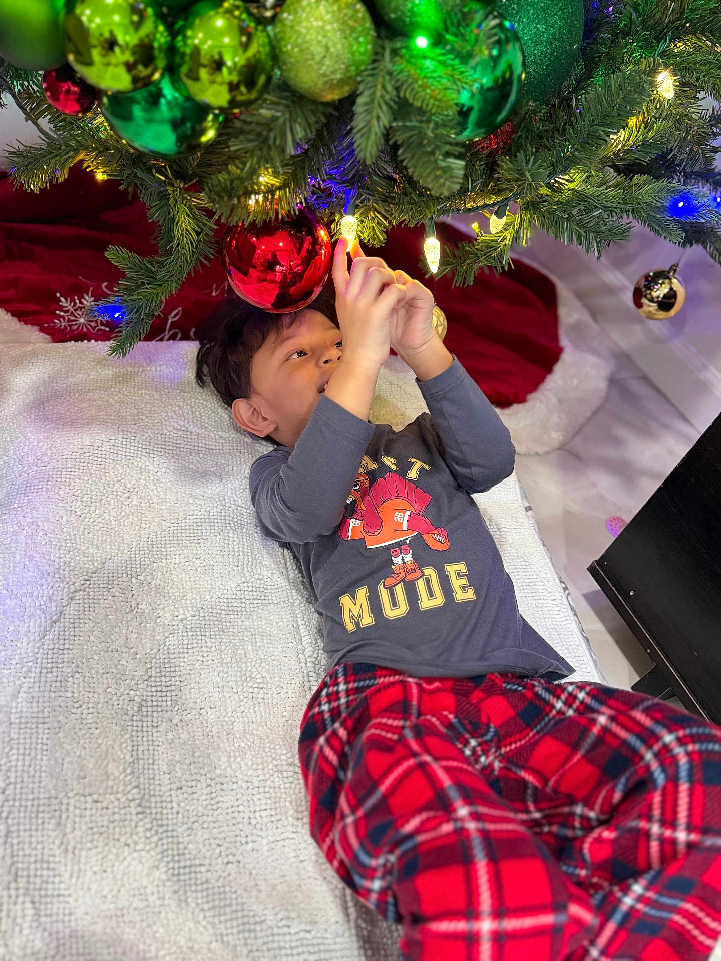 Ignacio joined the competition — help win amazing prizes! child, christmas_tree, christmas_ornaments, lights, blanket, red_pants, plaid, holiday, indoor, decorations, curious, reaching, festive, soft_texture, floor, playing, smiling, boy, cozy, holiday_season