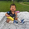 baby, banana, blanket, child, cute, grass, greenery, happy, jersey, outdoor, park, person, phone, play, sitting, smiling, sneakers, socks, toy, tree