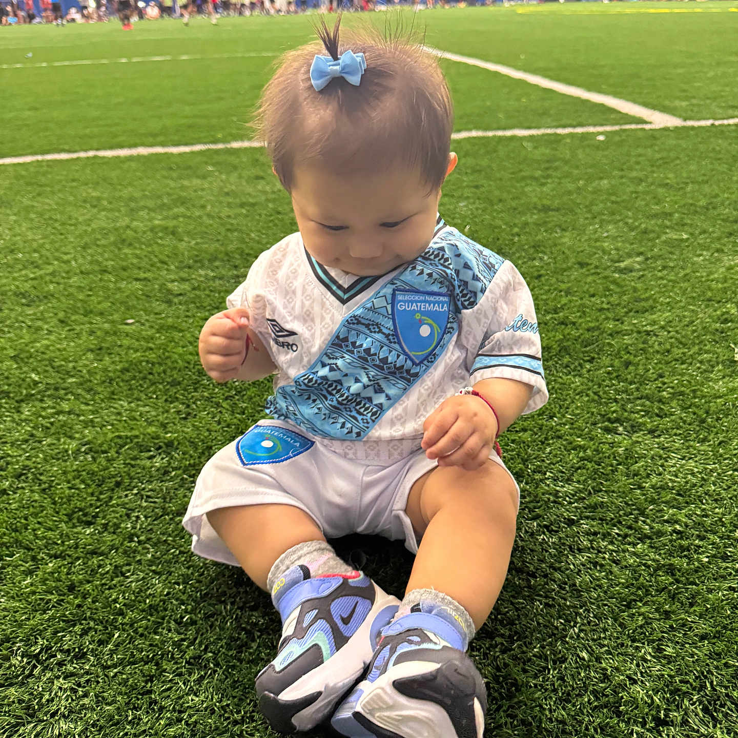 Keyri is registered to the contest to win money with this photo: baby, background, blue_bow, child, cute, field_lines, grass, green, guatemala, indoor, indoor_sports, jersey, people, playing, shoes, sitting, soccer_field, socks, sports, toddler
