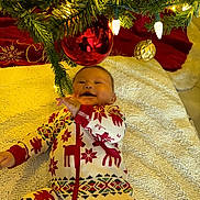 Keyri is registered to the contest to win money with this photo: baby, infant, christmas_tree, christmas_ornament, holiday, pajamas, festive_clothing, smiling, happy, indoor, blanket, decorations, red, green, lights, celebration, child, person, seasonal, festivity