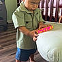 child, toddler, barefoot, toy, serious_expression, standing, wooden_floor, crib, chair, green_shirt, denim_shorts, indoor, furniture, person, home, concentration, short_hair, flooring, foot, hand