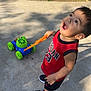 toddler, child, toy, mower, sidewalk, grass, red_jersey, shoes, outdoor, sunlight, curious, playing, summer, portrait, expression, young_child, casual_clothing, daytime, happy, person