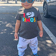 James is registered to the contest to win money with this photo: asphalt, car, casual, child, cute, daytime, dinosaur, fashion, happy, outdoor, pants, parking_lot, shadow, shirt, shoes, standing, sunglasses, sunlight, toddler, trees