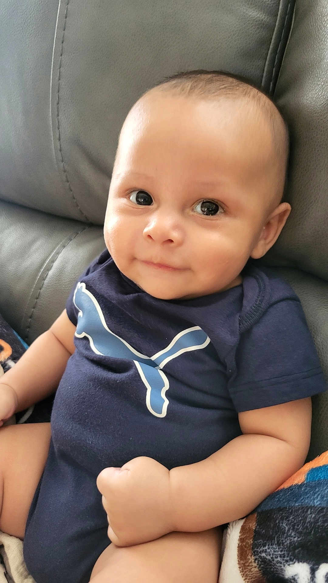 Ruben joined the competition — help win amazing prizes! baby, infant, smiling, onesie, blue_clothing, puma_logo, couch, leather_furniture, indoor, cute, child, portrait, sitting, chubby_cheeks, short_hair, skin, hand, arm, face, happy