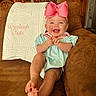 baby, pink_bow, smile, couch, quilt, embroidered_text, living_room, wooden_furniture, happy, child, indoor, foot, clothing, person, portrait, seat, home, infant, cute, cheerful