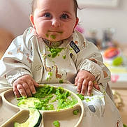 Arlo joined the competition — help win amazing prizes! baby, child, high_chair, bib, green_food, messy, feeding, cute, indoor, food, plate, hands, face, eyes, smile, clothing, person, infant, cucumber, meal