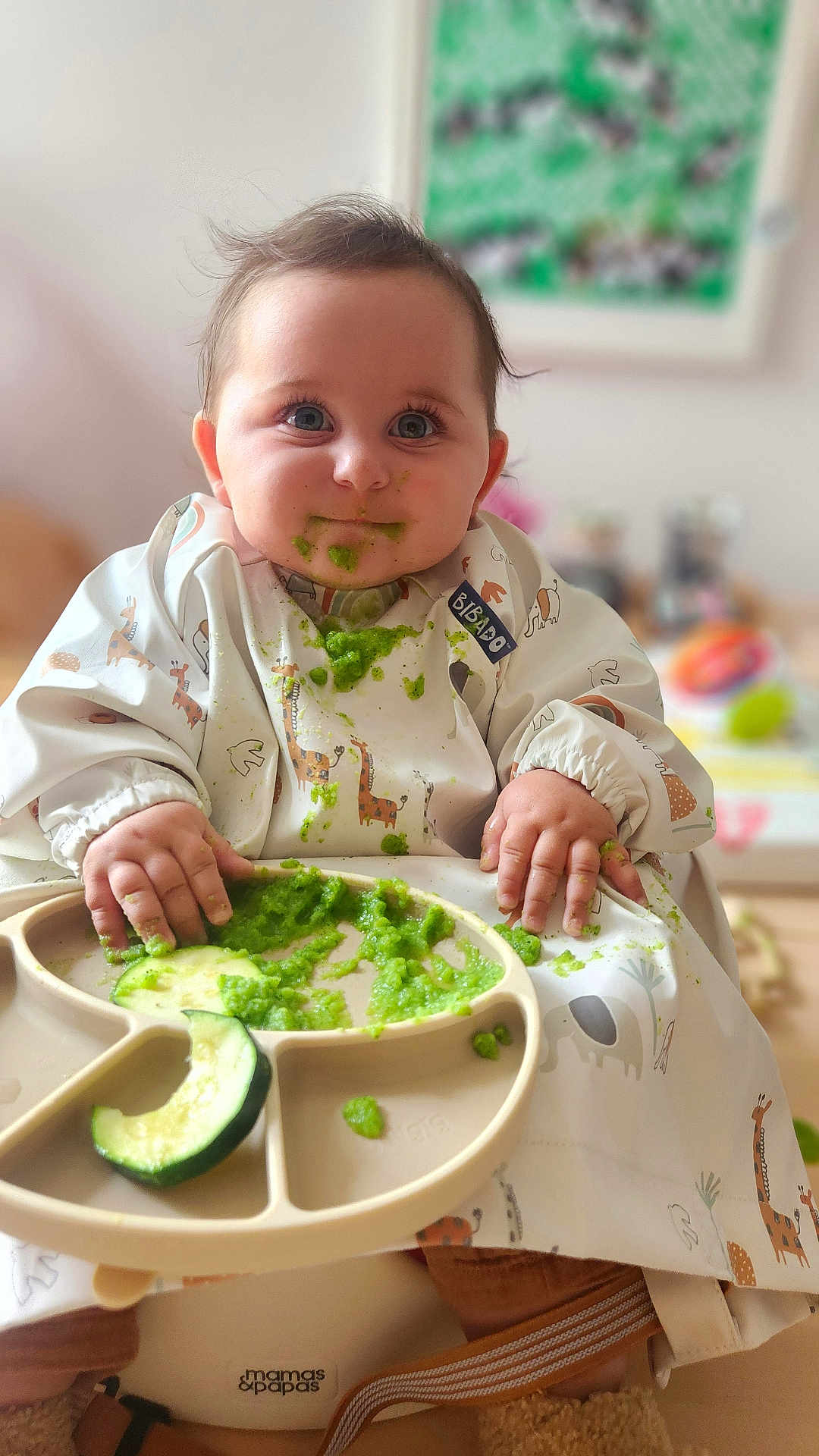 Arlo joined the competition — help win amazing prizes! baby, child, high_chair, bib, green_food, messy, feeding, cute, indoor, food, plate, hands, face, eyes, smile, clothing, person, infant, cucumber, meal